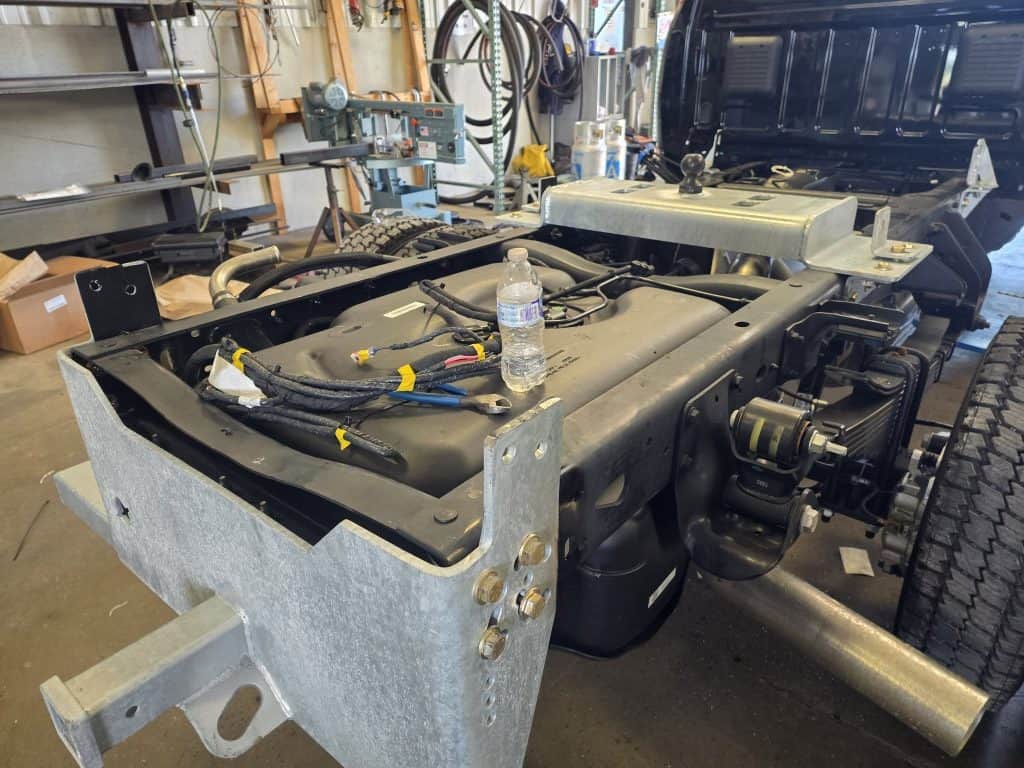 Work truck frame during professional flatbed installation showing wiring, gooseneck hitch mount, and secure mounting points in shop.